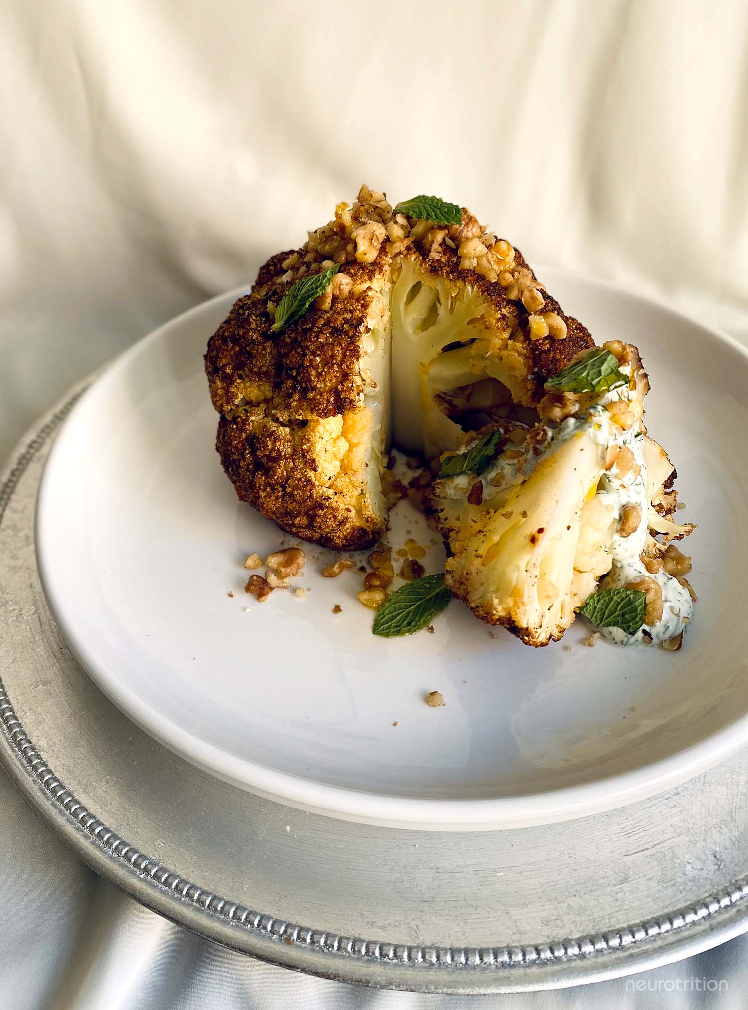 Whole roasted cauliflower with wedge cut out topped with herbs and yogurt sauce on a white plate