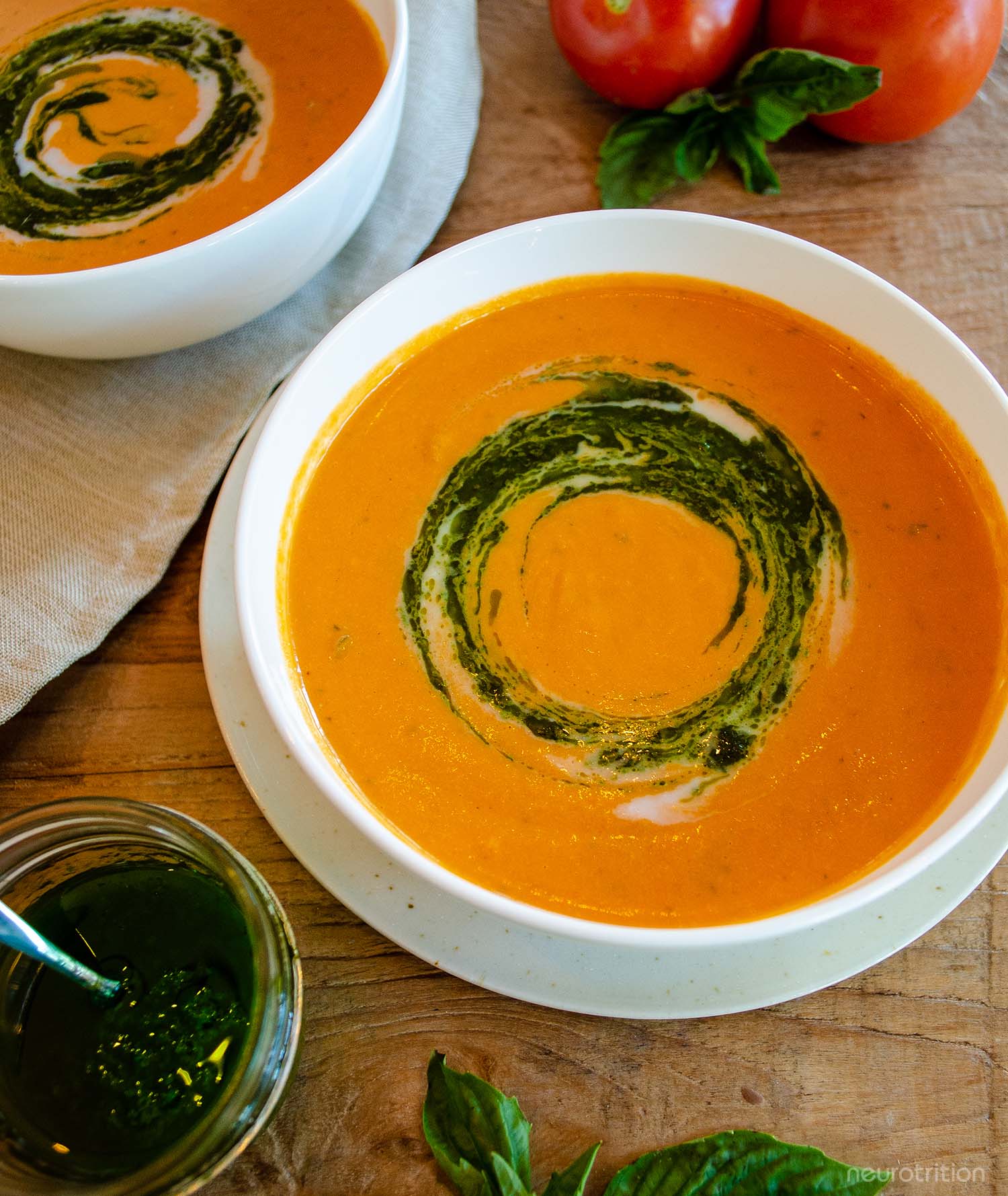 Two white bowls of roasted tomato soup sit on a wooden top table, with fresh whole tomatoes and a jar of pesto beside.