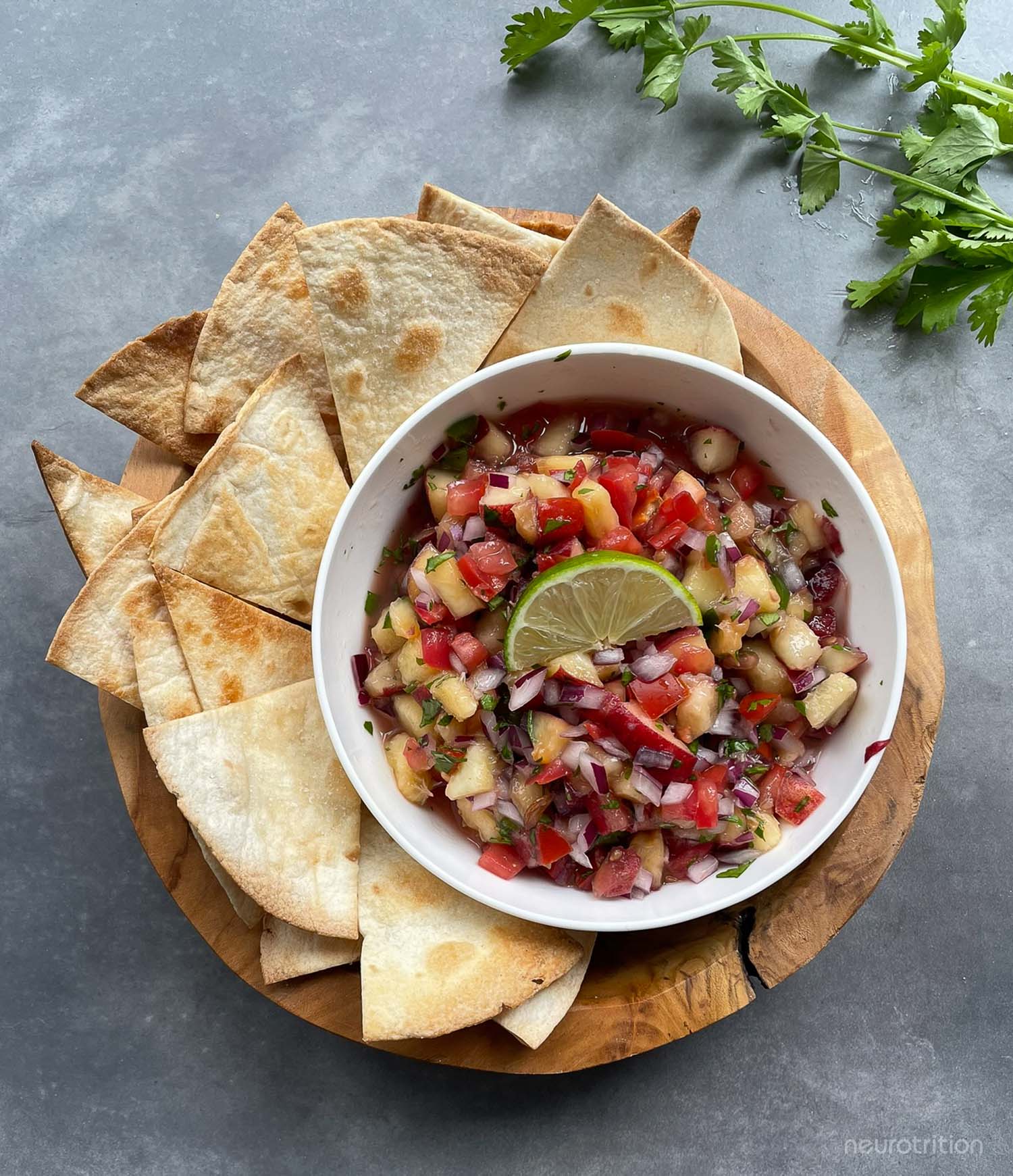 A bowl of homemade salsa is surrounded by baked pita chips on a wooden tray.