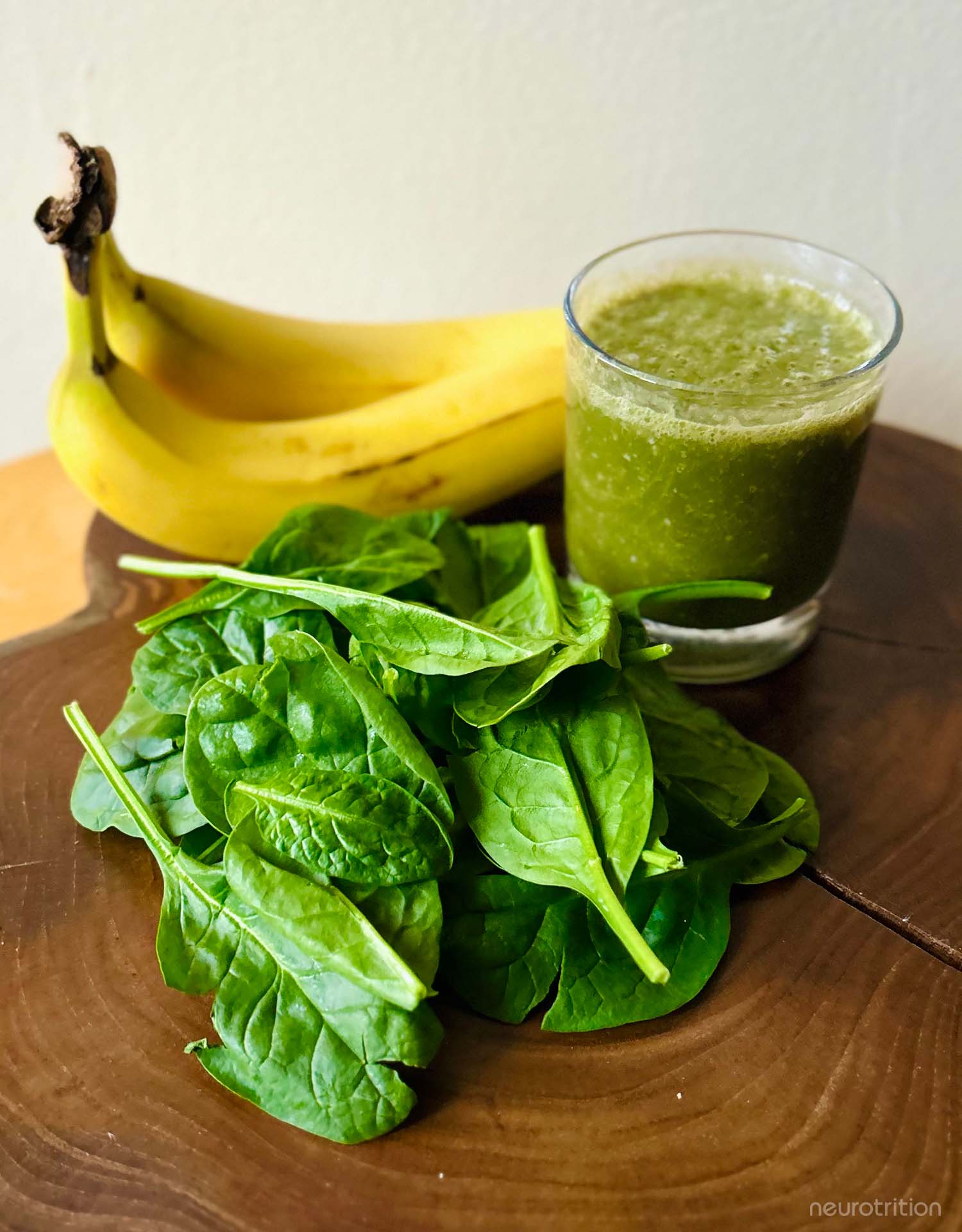 A green smoothie with bananas behind the glass and raw spinach sprinkled with hemp seeds in front, on a wooden board.