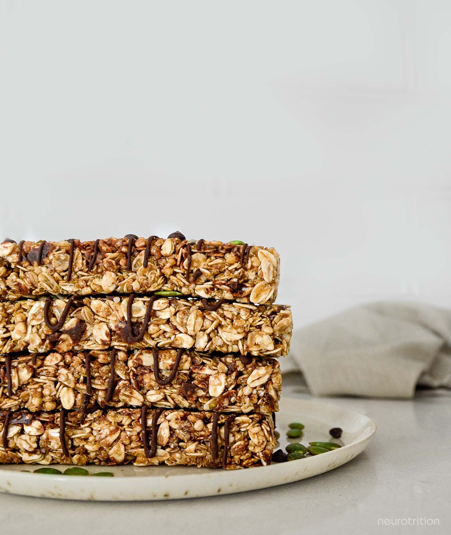 Four granola bars stacked on a beige plate, drizzled with chocolate and sprinkled with pumpkin seeds.