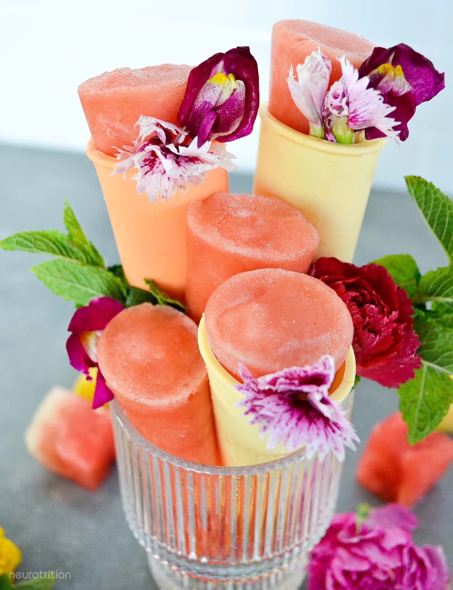 Five homemade pink popsicles in a glass, garnished with edible flowers and fresh mint.