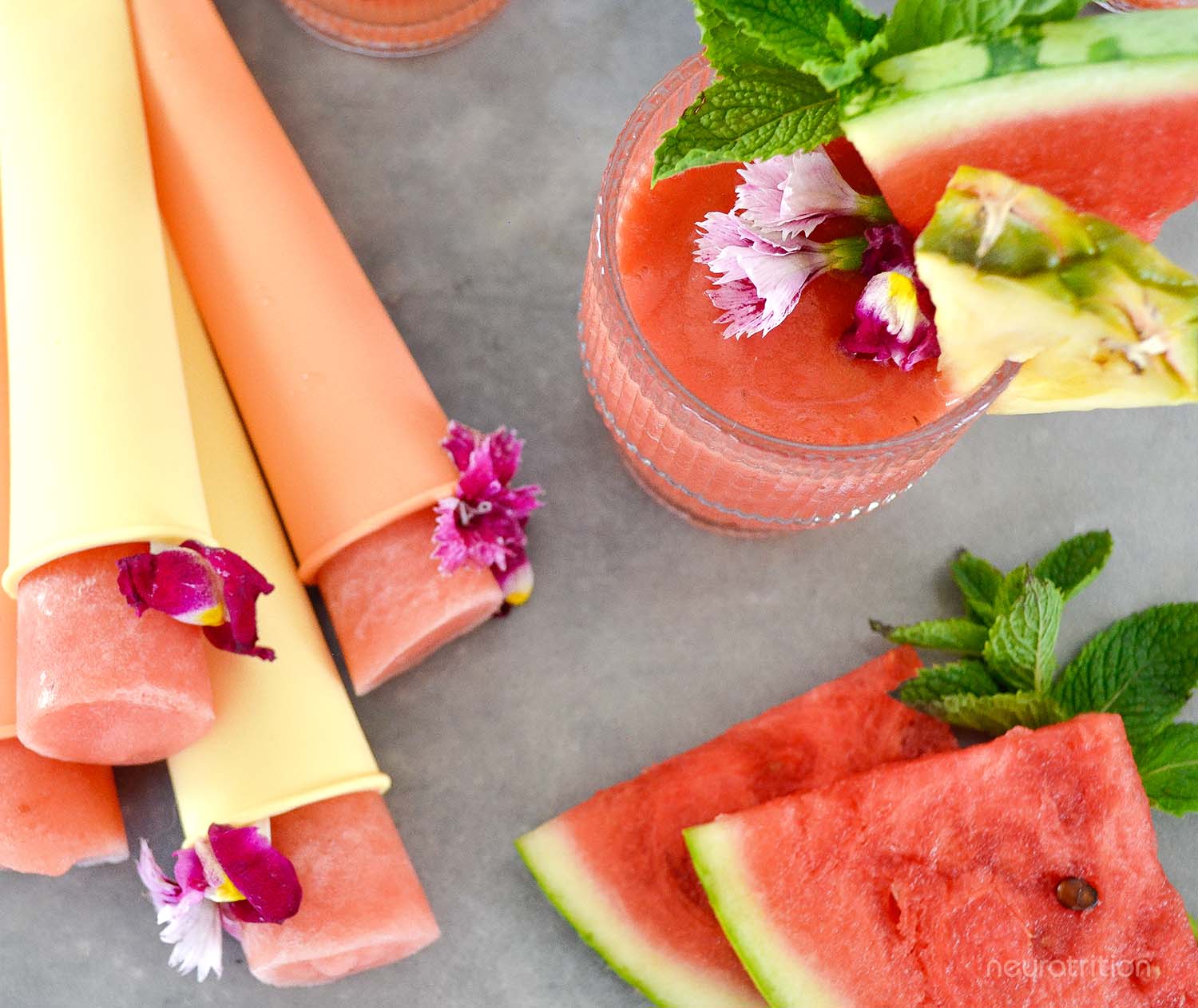 Top view of a watermelon pineapple coconut slushie in a glass with fruit and mint garnishes, beside homemade popsicles and slices of watermelon.