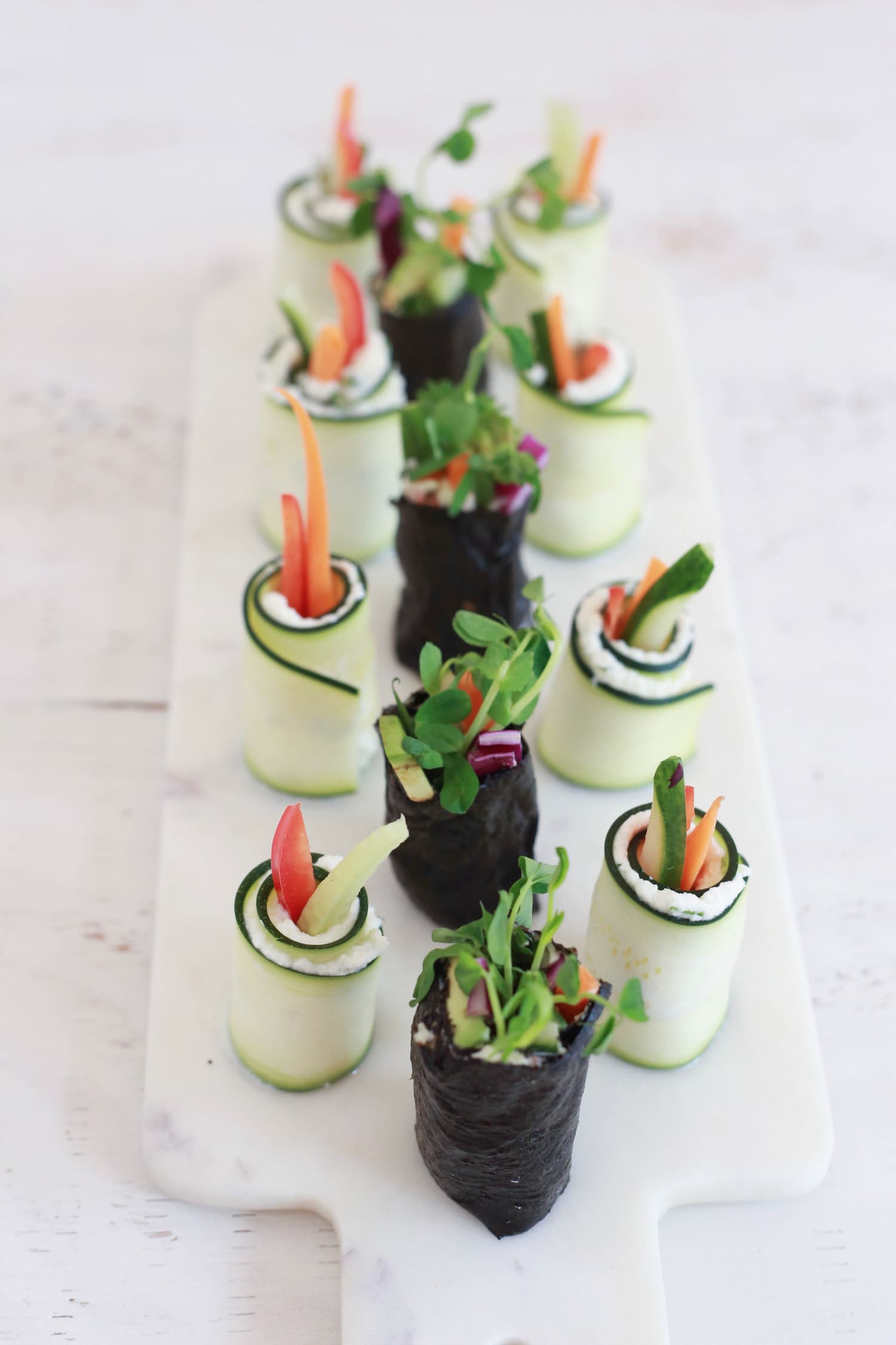 Veggie sushi wrapped in nori and raw zuchinni, on a white marble board.