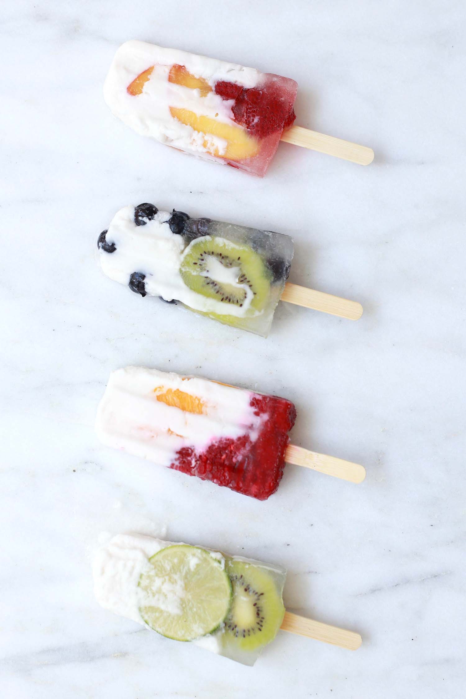 Four homemade popsicles in a row, with chunks of lime, blueberries, peaches and kiwi