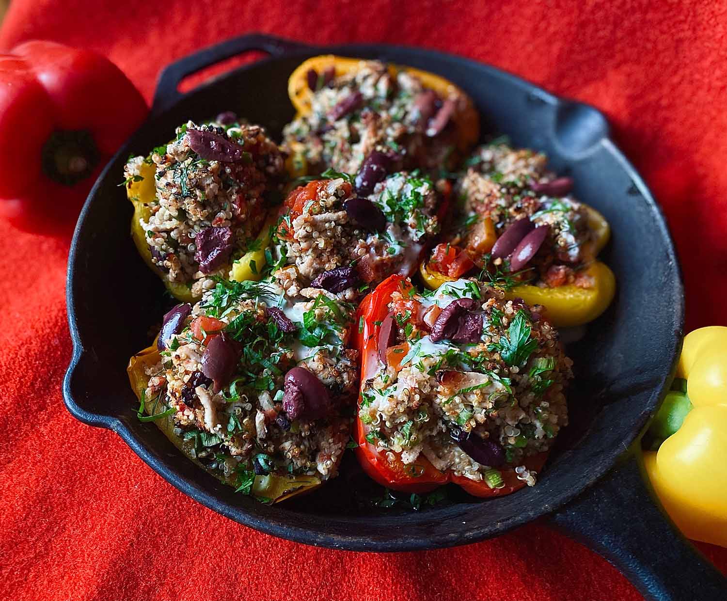 Red and yellow bell peppers stuffed with turkey, quinoa, olives, and herbs, in a cast iron pan, placed on a red linen background.