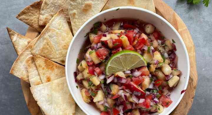 A bowl of homemade salsa is surrounded by baked pita chips on a wooden tray.