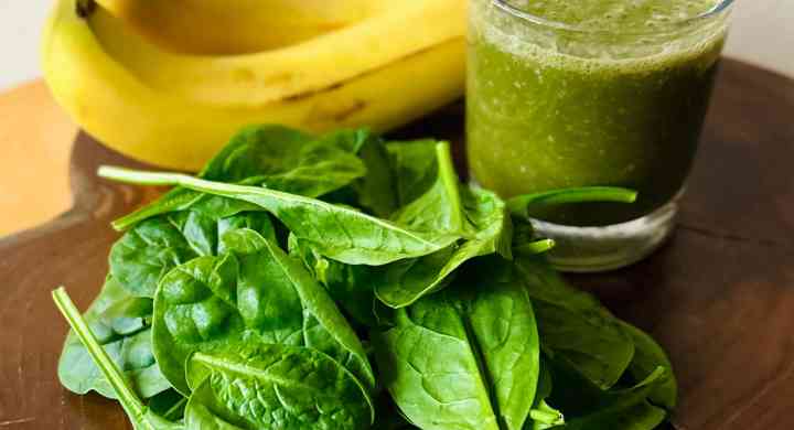 A green smoothie with bananas behind the glass and raw spinach sprinkled with hemp seeds in front, on a wooden board.