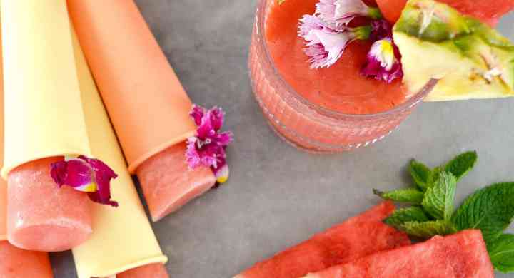 Top view of a watermelon pineapple coconut slushie in a glass with fruit and mint garnishes, beside homemade popsicles and slices of watermelon.