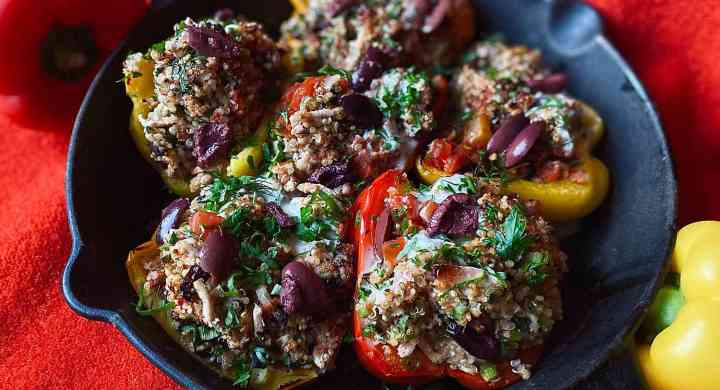 Red and yellow bell peppers stuffed with turkey, quinoa, olives, and herbs, in a cast iron pan, placed on a red linen background.