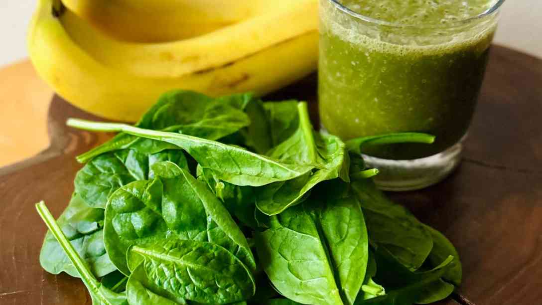A green smoothie with bananas behind the glass and raw spinach sprinkled with hemp seeds in front, on a wooden board.