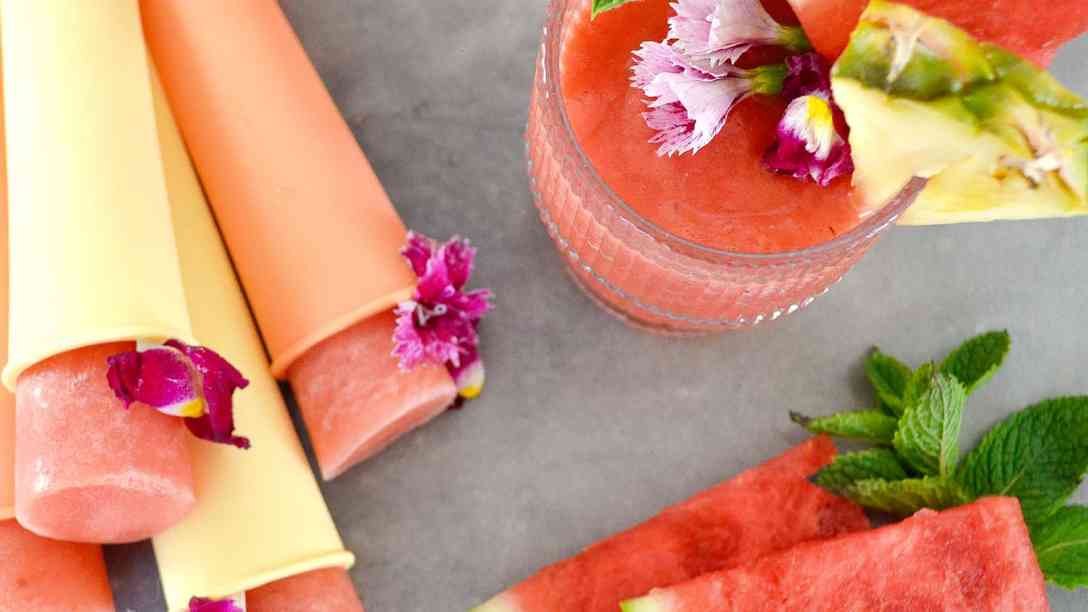 Top view of a watermelon pineapple coconut slushie in a glass with fruit and mint garnishes, beside homemade popsicles and slices of watermelon.
