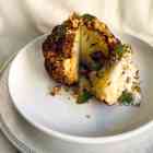 Whole roasted cauliflower with wedge cut out topped with herbs and yogurt sauce on a white plate