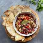 A bowl of homemade salsa is surrounded by baked pita chips on a wooden tray.